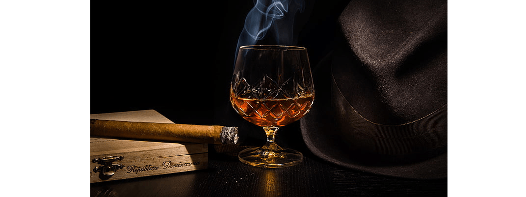 A smoking cigar on top of a wooden humidor, with a bit of ash on the end. A glass of brandy, and a brown fedora.